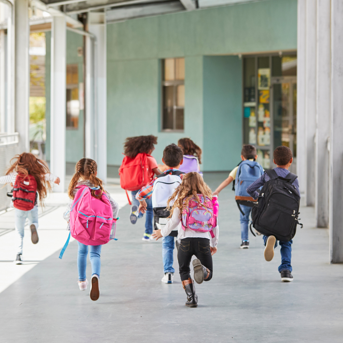 A group of children running toward a school building with backpacks, seen from behind.
