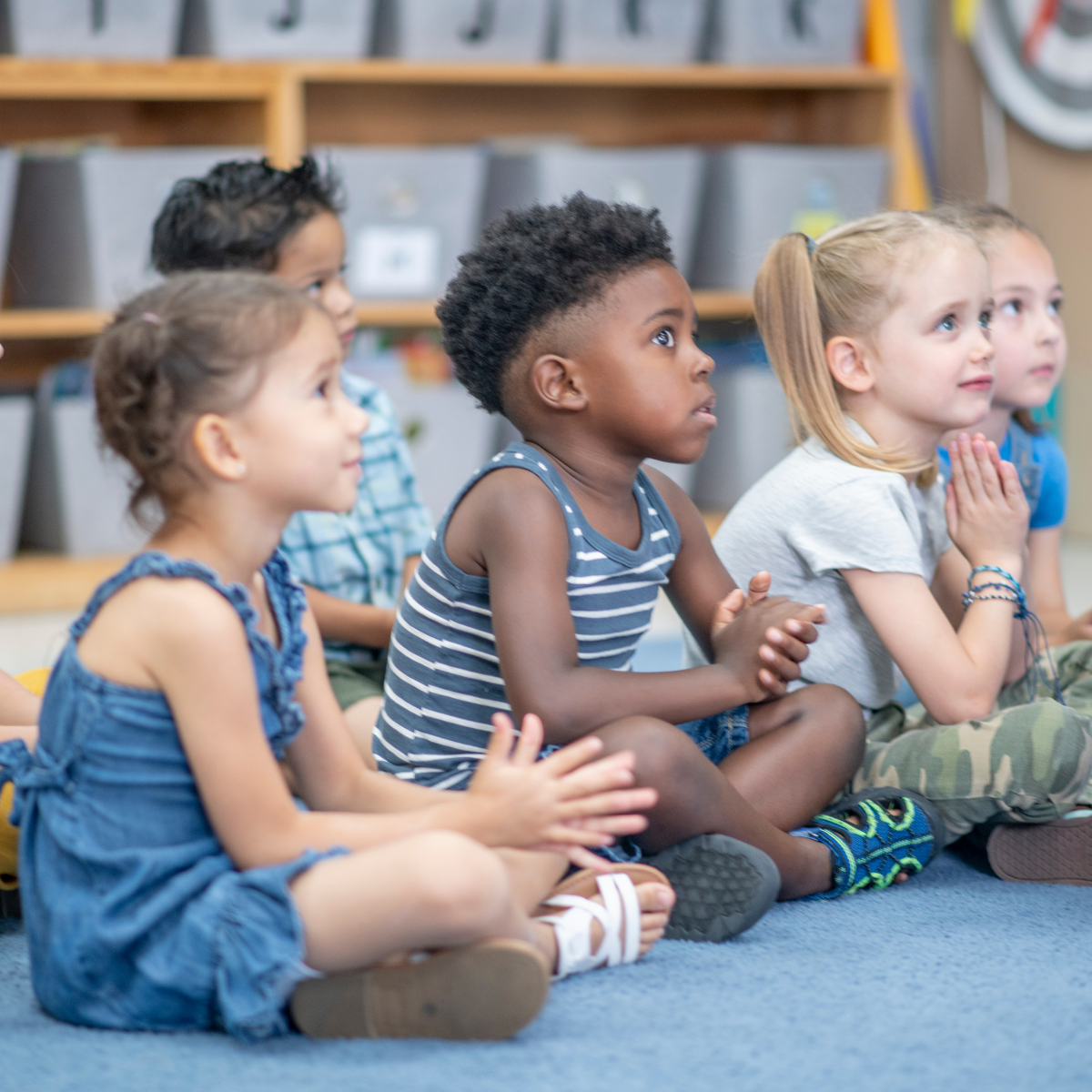 A row of young students seated on the classroom floor, in profile, listening intently.