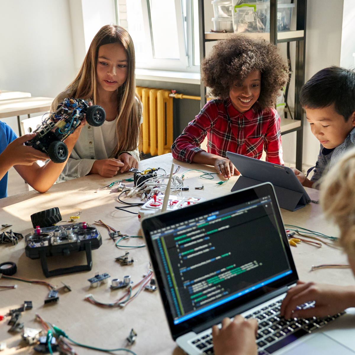 Students at a maker table with a laptop showing code, robot parts and wires spread across the workspace.